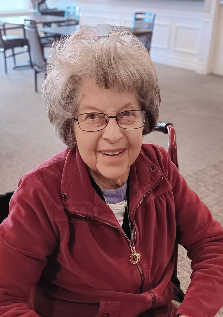The image of May is an older woman sitting in the community, wearing a red jacket and glasses.