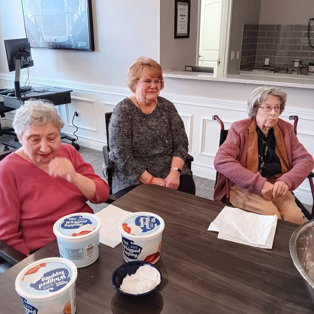 Three elderly women and volunteers gather around a table, preparing desserts with whipped topping