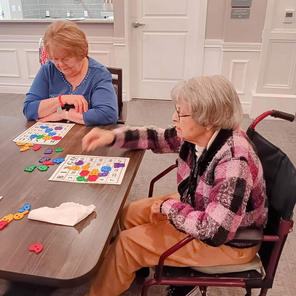Two joyful elderly women engage in a lively game of cards, surrounded by caring volunteers in a memory care neighborhood.