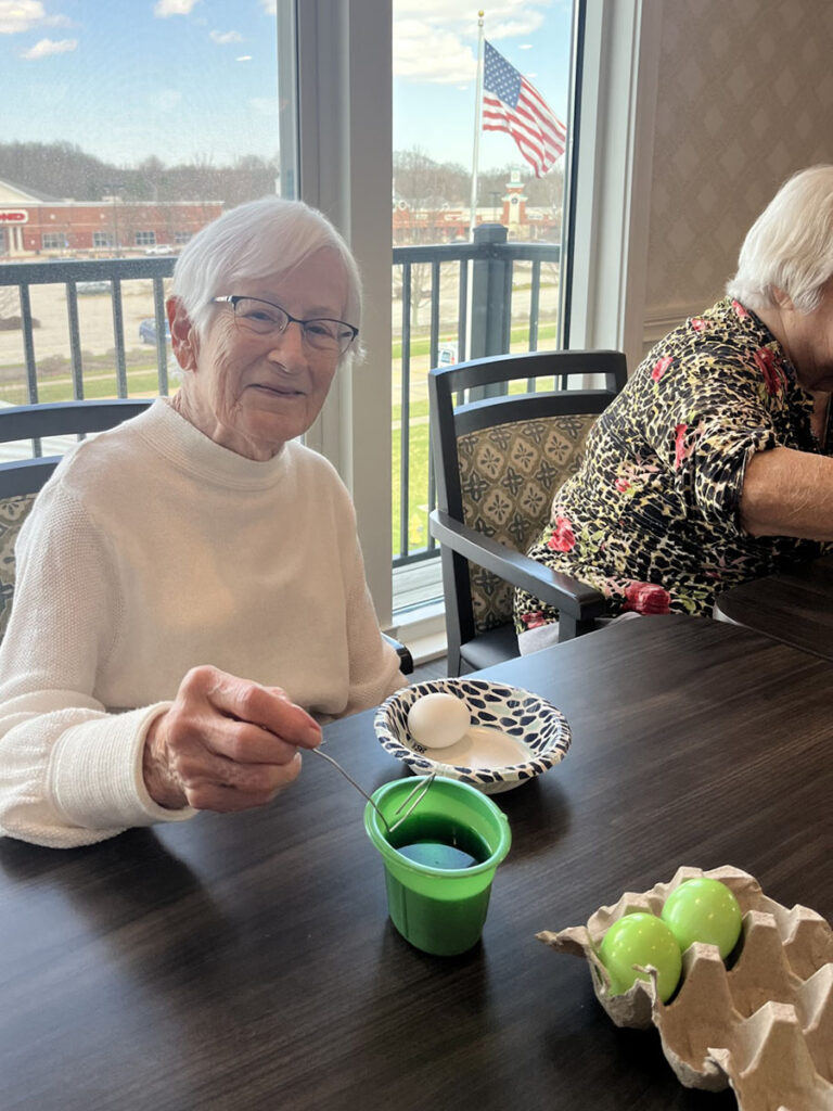 Two elderly women sit at a table with eggs and a cup. One senior living resident smiles while dyeing an Easter egg green.