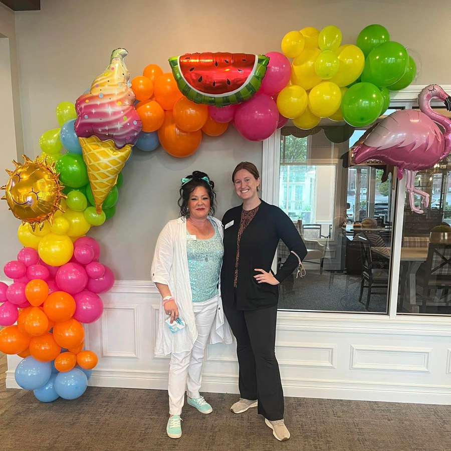 Two women stand smiling under a colorful balloon arch decorated with summer-themed inflatables, including an ice cream cone, watermelon slice, sun, and flamingo.