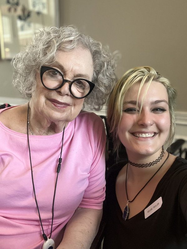 A senior resident and a staff member smile together for a selfie. The senior wears glasses and a pink top, while the staff member has short hair and a choker.