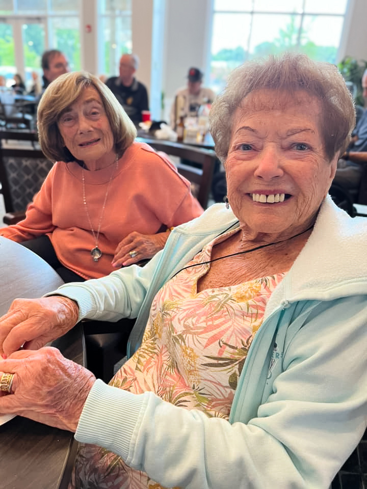 Two smiling senior residents sit at a table during a social gathering. One wears a floral dress and light jacket, and the other is dressed in a peach-colored top.