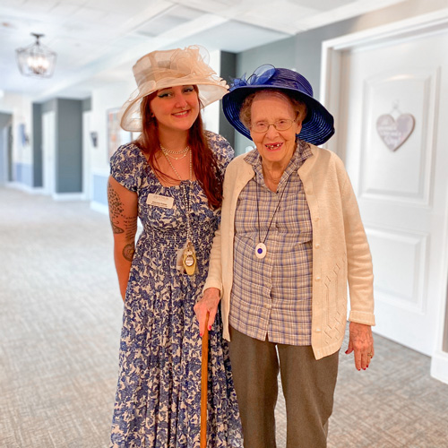 Senior resident and team member at a senior living community dressed in elegant outfits with hats, standing together in a well-lit hallway, sharing smiles during a special event.