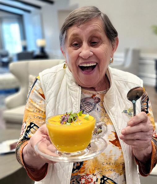Smiling senior resident joyfully holds a bowl of vibrant orange soup garnished with greens, wearing a colorful butterfly-patterned shirt and white vest, ready to enjoy the meal.