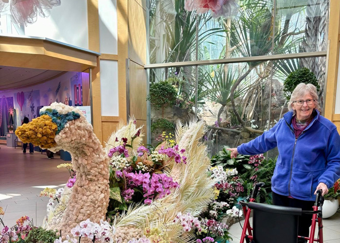 Senior woman smiling next to a large floral swan sculpture at a botanical garden.