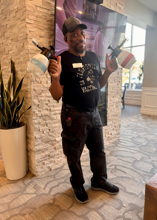 Maintenance team member holding two cleaning solution sprayers while smiling in a senior living lobby.