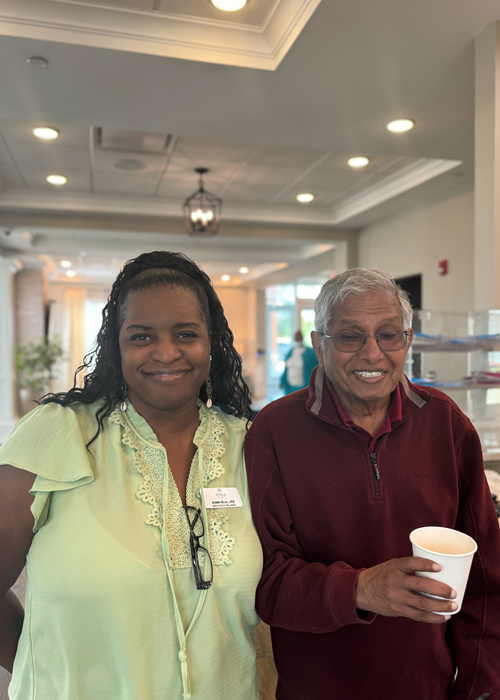 Team member in a light green blouse stands next to a smiling senior resident in a burgundy sweater holding a white cup, both enjoying time together in the bright, welcoming lobby of a senior living community.