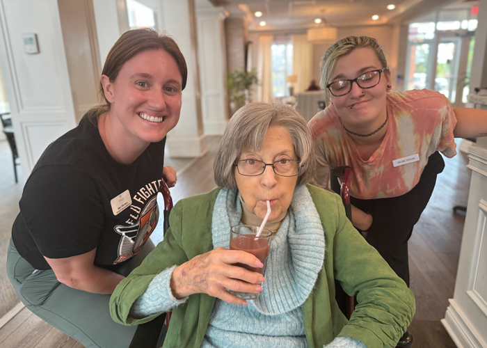 Senior resident sips a red drink through a straw while seated between two smiling team members, creating a joyful and engaging moment inside a senior living dining area.
