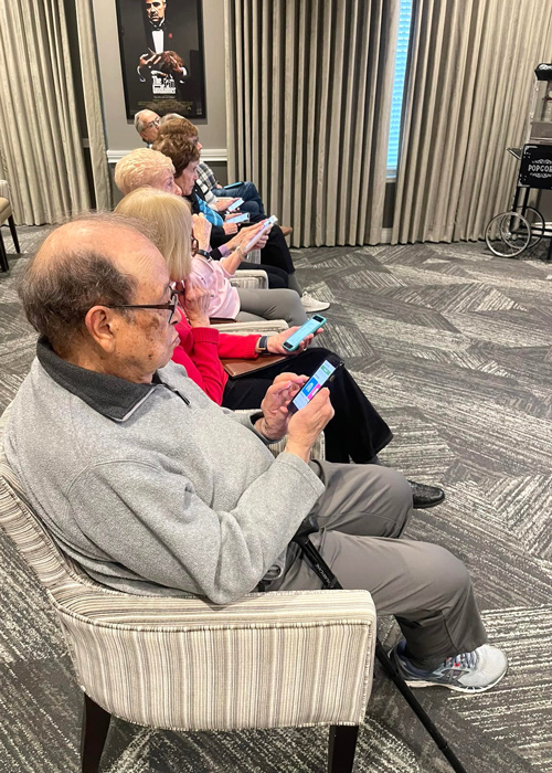 Senior residents sit in a row, each engaged with their smartphones during a technology learning session in a comfortable community room.