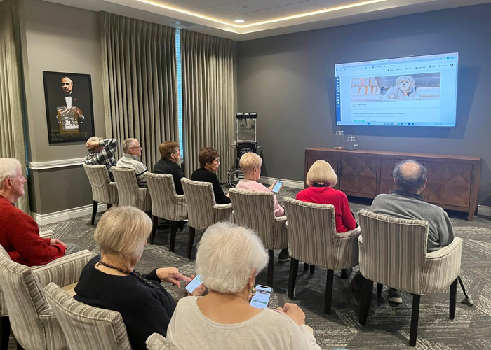 A group of senior residents attentively follows a technology training presentation displayed on a large screen, many using their phones to participate.