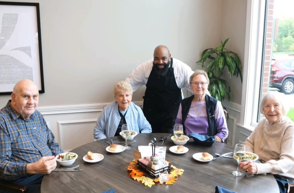 A Culinary Director smiles with four senior residents around a dining table, enjoying fresh salads, rolls, and wine in a bright, welcoming community setting.