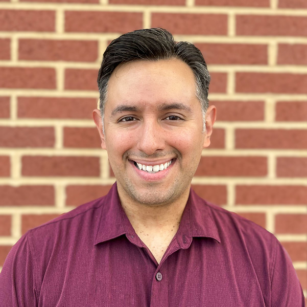 Adam Rodriguez, Resident Services Director at Vitalia Solon, smiling in a professional headshot. He has short dark hair, wears a burgundy button-down shirt, and is standing in front of a red brick wall.