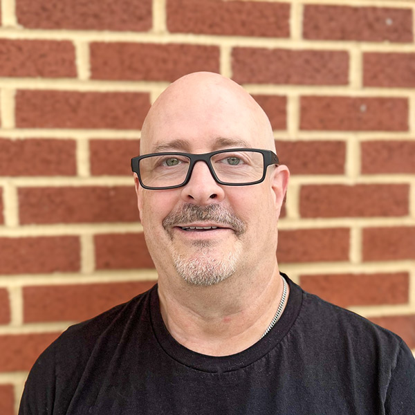 David Granito, Wellness Director at Vitalia Solon, smiling in a professional headshot. He is bald with a goatee, wears black rectangular glasses and a dark shirt, standing in front of a red brick wall.