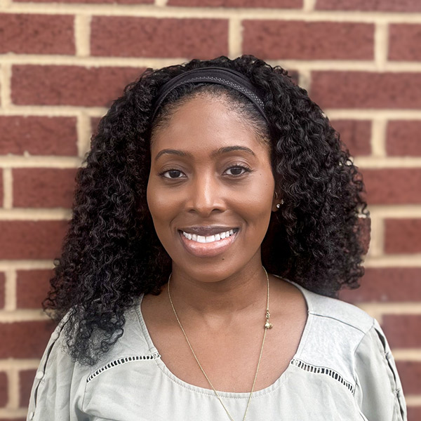 Erika Threat, Assistant Executive Director at Vitalia Solon, smiling in a professional headshot. She has long curly hair held back with a black headband and wears a light blouse with a delicate necklace, standing in front of a brick wall.