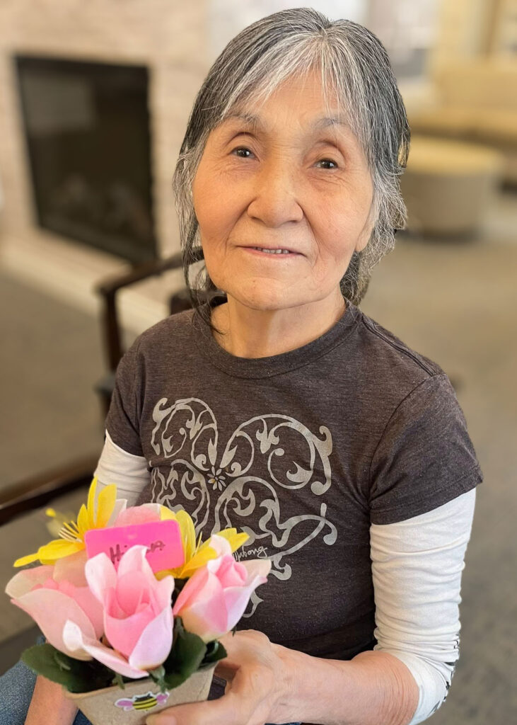A smiling senior woman proudly holds a bouquet of handmade pink and yellow paper flowers, showcasing her craft project in a warm and inviting community setting.