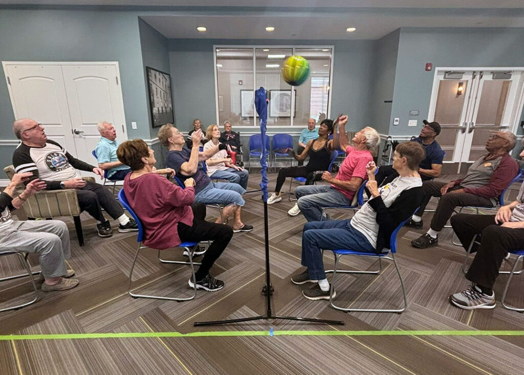 A group of residents and team members play a game of chair beachball volleyball in the fitness center.