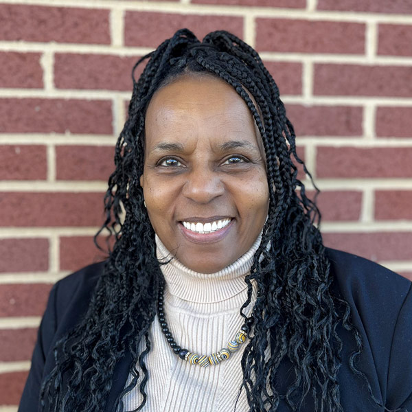 Annette Dendy, Resident Services Director at Vitalia Solon, smiling in front of a brick wall. She has long braided hair, wears a beige turtleneck with a beaded necklace, and a black blazer.