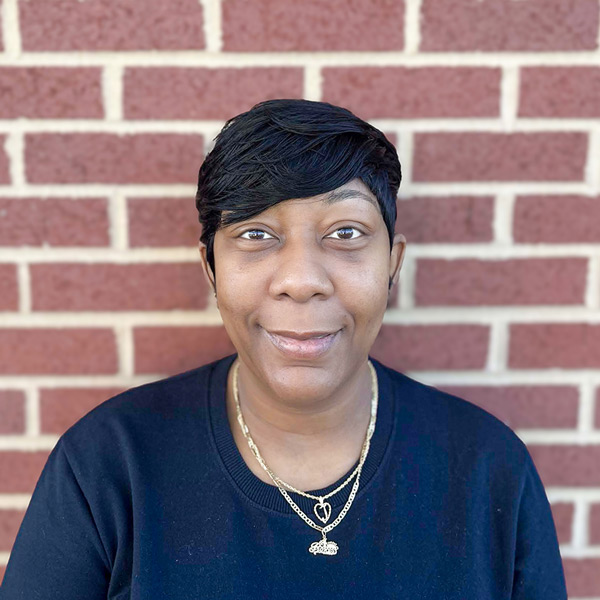 Jocelyn Zellous, Memory Care Director at Vitalia Solon, smiling in front of a red brick wall. She is wearing a black top and gold layered necklaces with heart and pendant charms.