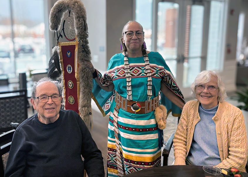 Senior residents sit smiling beside a presenter wearing traditional Native American regalia, holding a ceremonial item during a cultural education event.