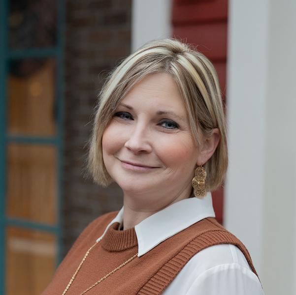 Ann Mills, Memory Care Director at Vitalia Solon, smiling in a professional headshot, wearing a brown sweater vest over a white blouse, standing outdoors with a softly blurred building background.