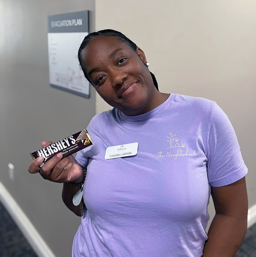 A team member wearing a purple shirt smiles, holding a sweet treat.