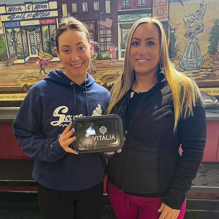 A team member from Vitalia Solon smiles with a woman at a local business, standing in front of a colorful mural, with a gift from the community.