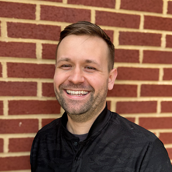 Jonathan “Jon” Blaha, Wellness Director at Vitalia Solon, smiling in a professional headshot with short light brown hair, light stubble, a black collared top, and a red brick wall background.