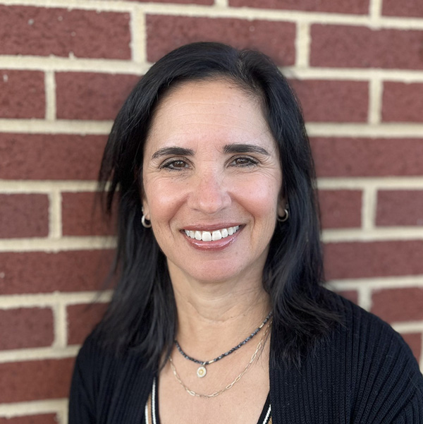 Sheryl Chaitoff, Senior Living Coordinator at Vitalia Solon, smiling in a professional headshot with straight dark shoulder-length hair, hoop earrings, layered necklaces, and a red brick wall background.