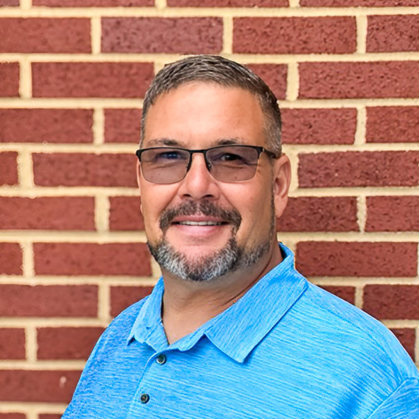 Mark Beran, Plant Operations Director at Vitalia Solon, smiling in a professional headshot with short gray hair, glasses, a salt-and-pepper goatee, a bright blue polo shirt, and a red brick wall background.
