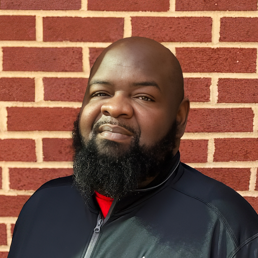 Charles Garner, Culinary Director at Vitalia Solon, smiling in a professional headshot, wearing a black zip-up jacket over a red shirt, posed in front of a red brick wall.