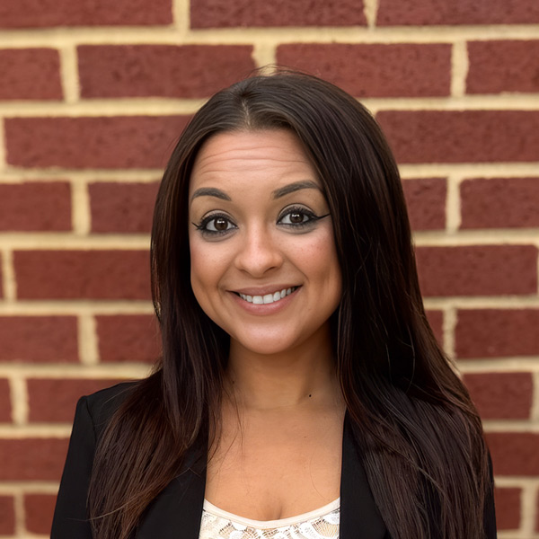 Paige Moltzan, Senior Living Director at Vitalia Solon, smiling in a professional headshot with long dark brown hair, a black blazer, and a red brick wall background.